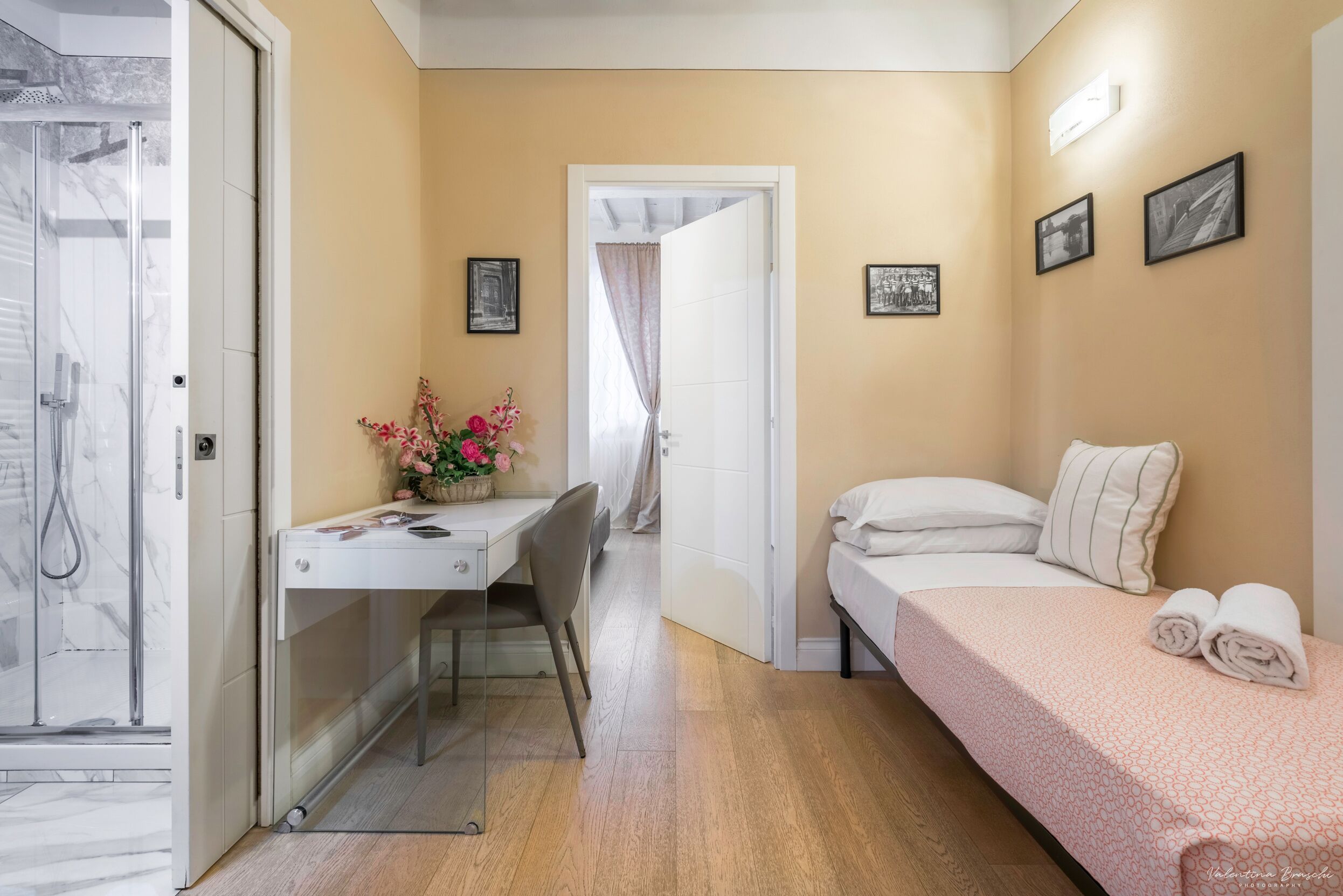 Habitación de hotel pequeña con ducha, un escritorio y una silla, cama con almohadas y una mesita con flores.