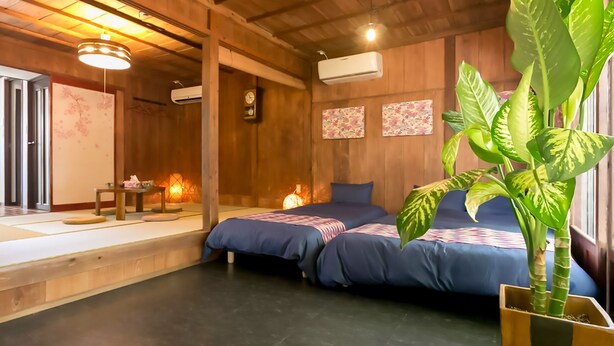 A traditional Japanese-style room with tatami flooring, two beds with blue bedding, a wooden ceiling, and a large green plant.