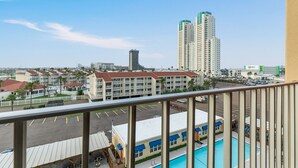 A view from a balcony overlooking a pool area with a parking lot, a hotel building, and high-rise buildings in the background.