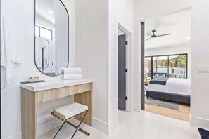 A modern bathroom with a large mirror, white vanity, and a small stool. A bedroom with a bed and a view of the outdoors is visible through an open door.