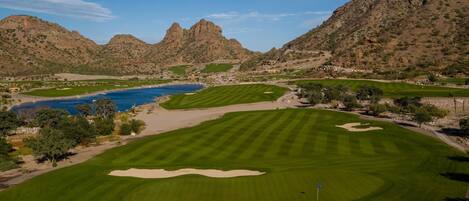 A golf course with a water body, sand bunkers, and mountainous terrain.
