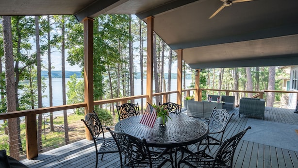 Une terrasse extérieure couverte, dotée d’une table ronde et de chaises, qui offre une vue sur un lac et est entourée d’arbres.
