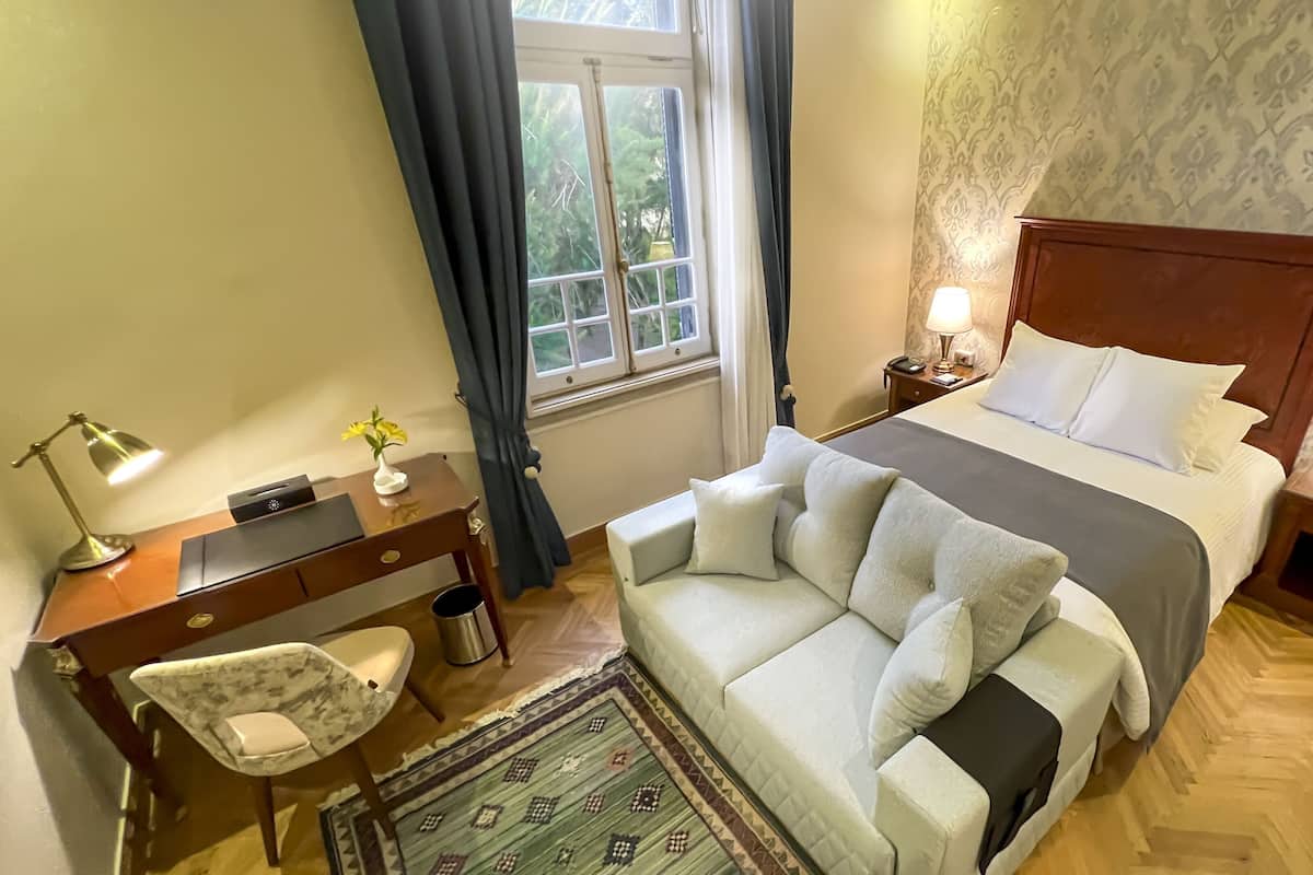 Ein Hotelzimmer mit einem Bett, einem Schreibtisch mit Stuhl, einer Lampe, einem kleinen Tisch mit einer Vase und einem Fenster mit Vorhängen.