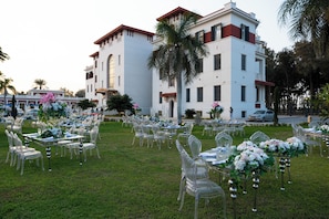 An outdoor wedding venue with clear glass tables and chairs, a white building in the background, and a well-maintained lawn. - The Palace Hotel - Adults Only (Alexandria)
