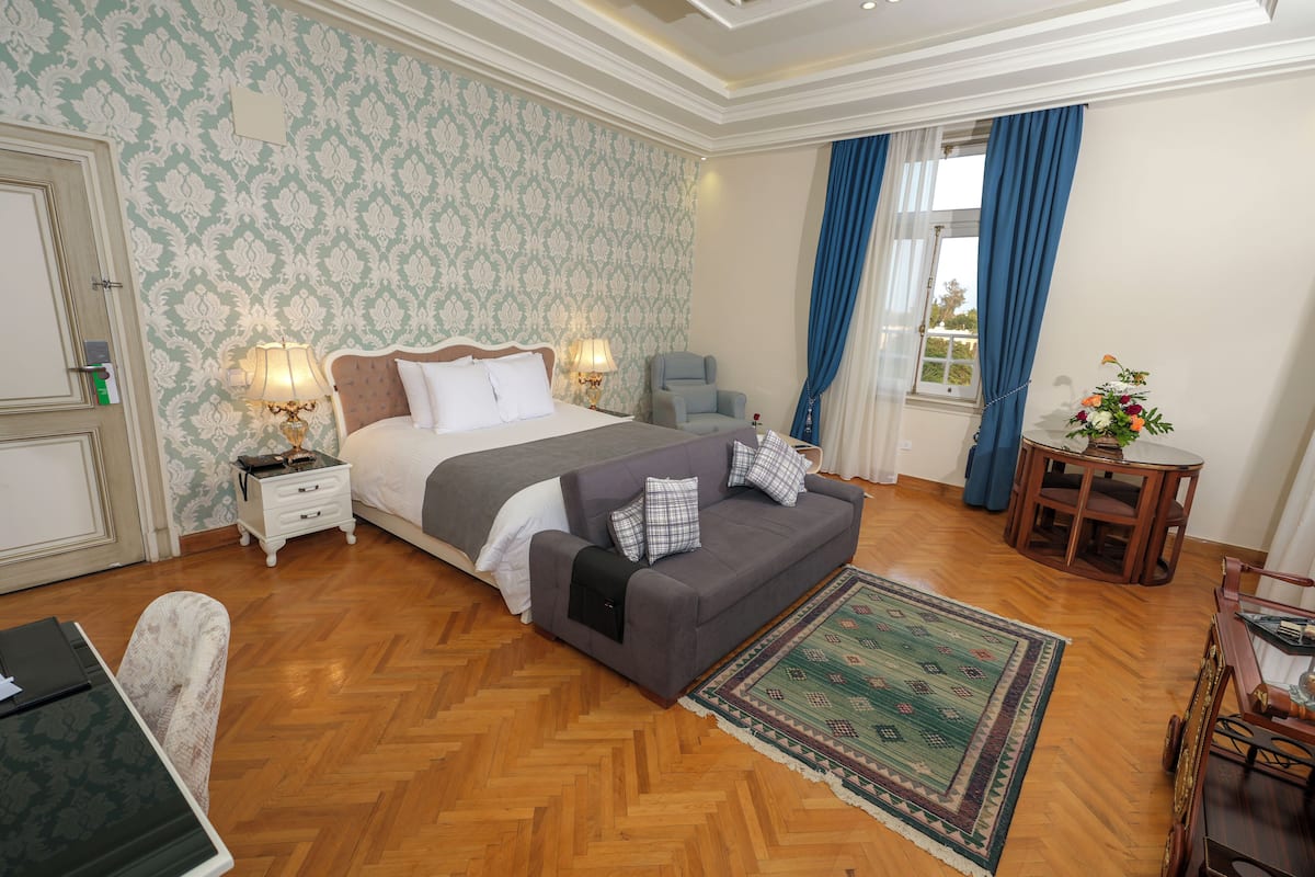Ein Schlafzimmer mit Bett, Nachttischen, einem Sofa, einem kleinen Tisch mit einer Vase und einem Fenster mit Vorhängen.