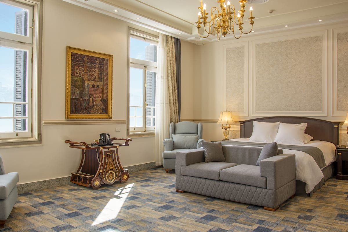 Ein Hotelzimmer mit einem großen Bett, einer Couch, einem Sessel, einem kleinen Tisch mit Lampe und einem Gemälde an der Wand.
