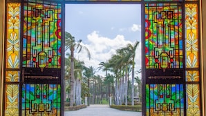 Une vue à travers un vitrail aux motifs géométriques et aux thèmes tropicaux, qui donne sur un sentier bordé de palmiers.