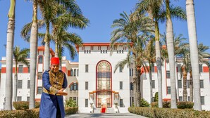 Un homme, vêtu d’un costume traditionnel, se tient devant un bâtiment dont le toit est recouvert de tuiles rouges, entouré de palmiers et dont un escalier mène à l’entrée.