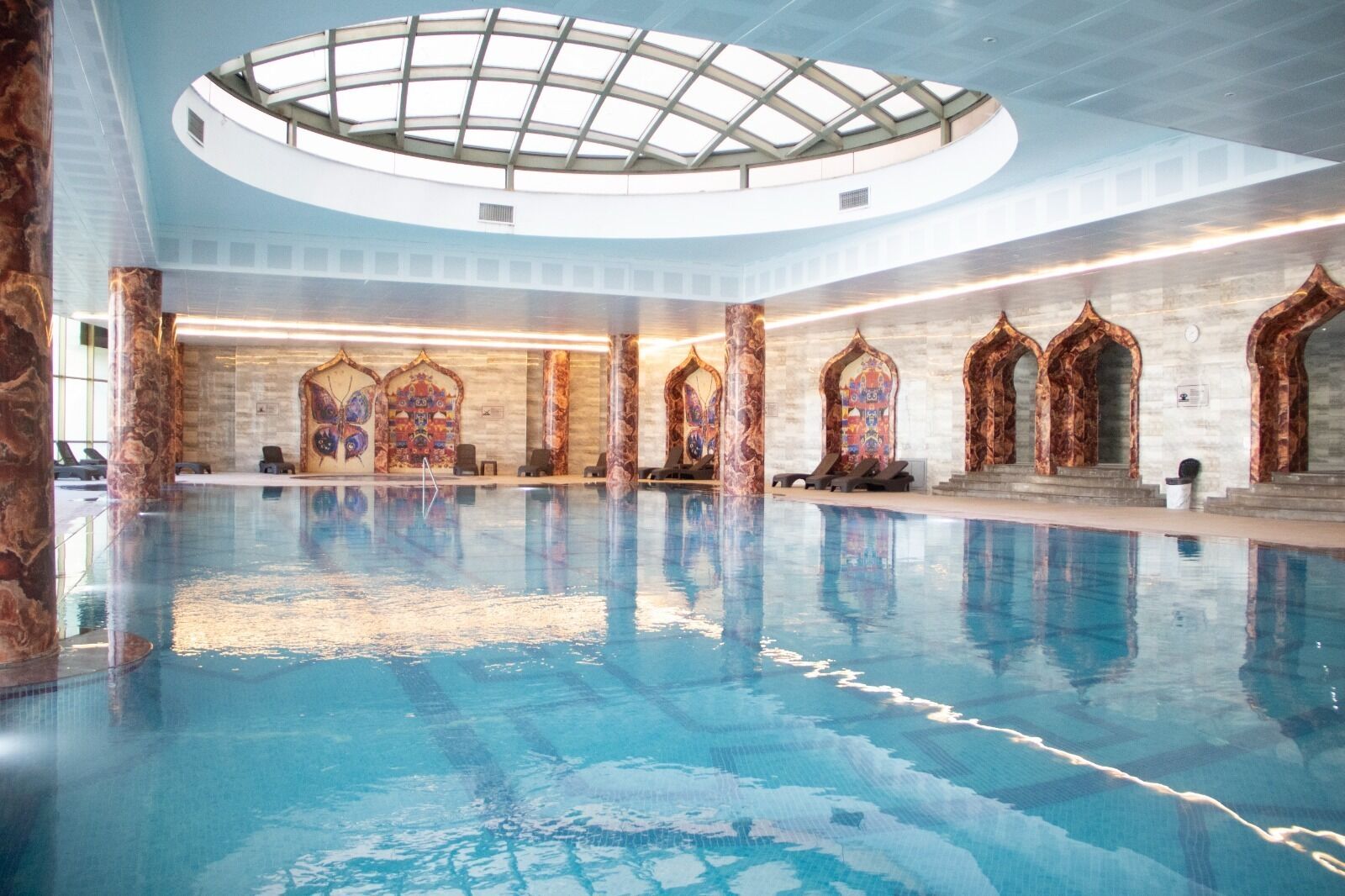 An indoor swimming pool with a glass ceiling, marble columns, and ornate wall decorations.