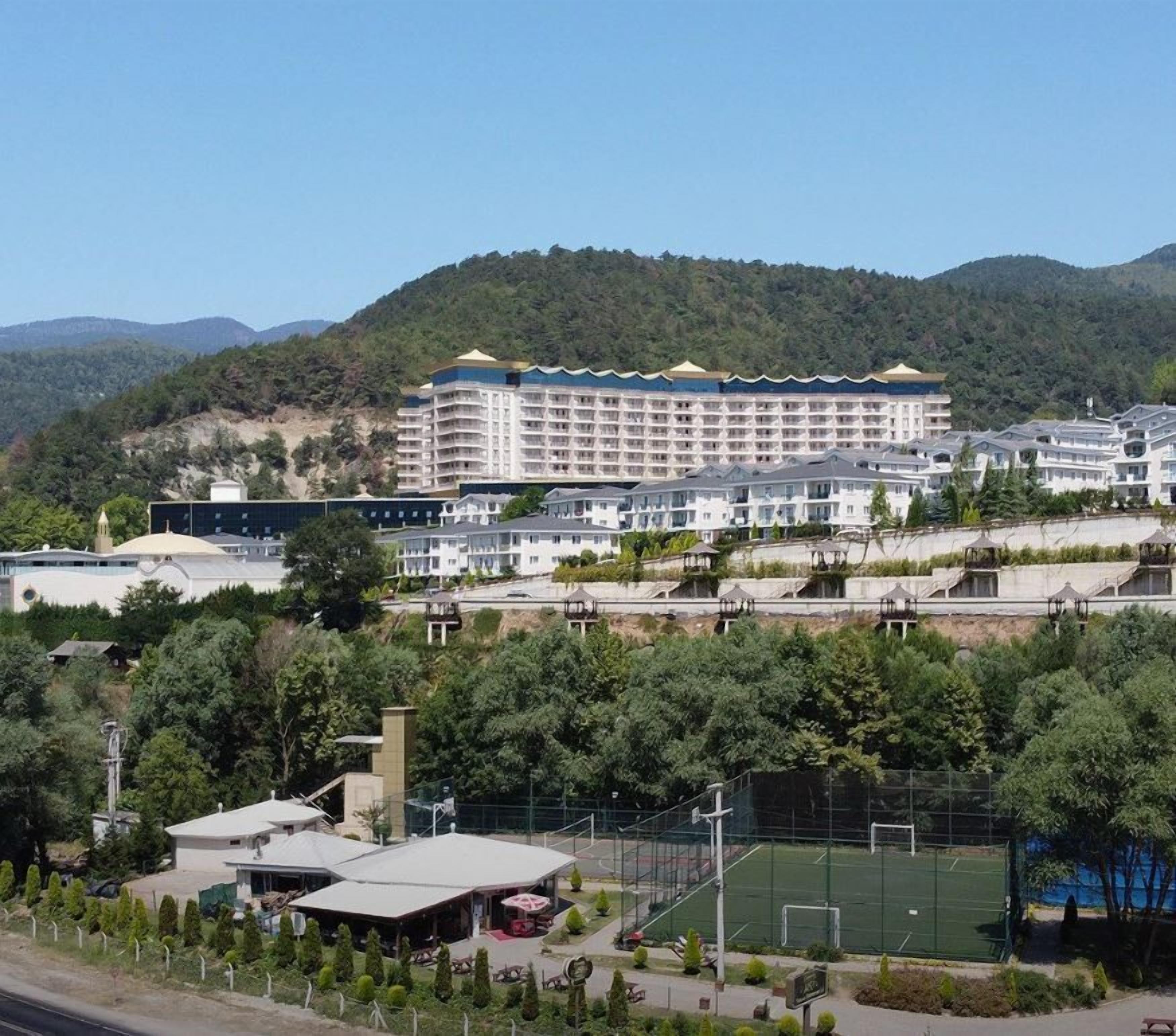 Ein mehrstöckiges Hotel, ein großes Gebäude mit flachem Dach und eine umliegende Grünfläche mit Bäumen und einem Tennisplatz.