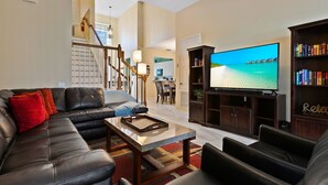 A living room with a leather sofa, a coffee table, and a flat-screen TV displaying a beach scene. There is a staircase leading to another room, and a bookshelf filled with books. - Last Minute Disney Getaway! Private Pool, Hot Tub & Game Room. Book Now! #1388 (Orlando)