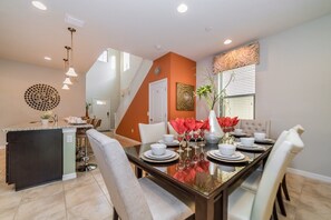 A modern dining area with a glass-top table, white chairs, and a central vase with red flowers. - Last Minute Disney! Private Pool, Hot Tub, Game Room & Themed Bedrooms. Book Now! #15098 (Orlando)