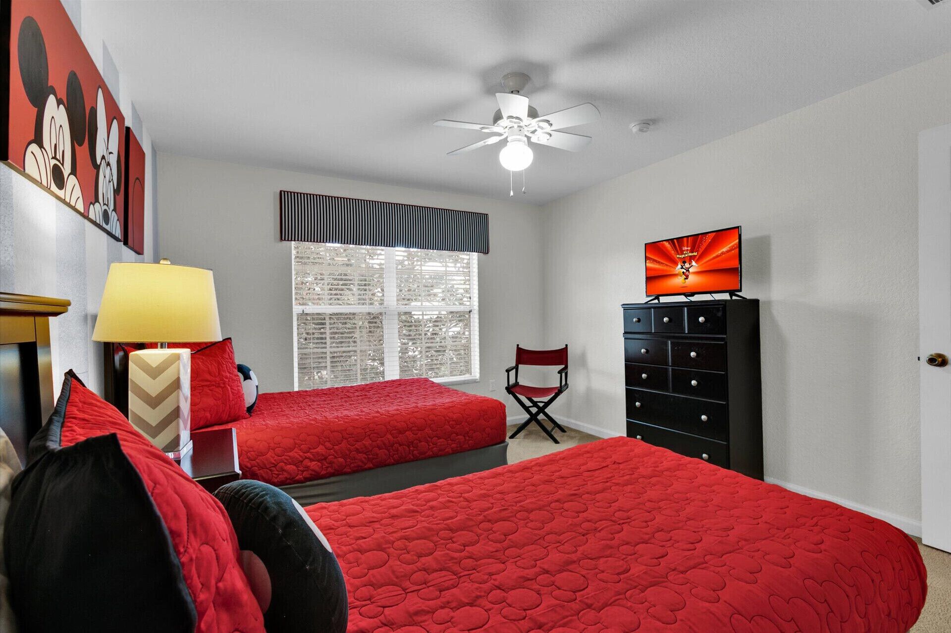 A bedroom with two beds, a ceiling fan, a window with blinds, a TV on a dresser, and a chair.