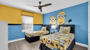 A bedroom with two beds, a ceiling fan, a window with blinds, and a wall mural of Minions.