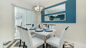 Un coin repas avec une table blanche, des sets de table bleus, des chaises blanches, un lustre et un mur d’accent bleu.