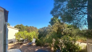 Un espace extérieur aménagé en patio, avec une table ronde et des chaises, un fauteuil de relaxation et un petit jardin agrémenté d’arbres et d’arbustes.