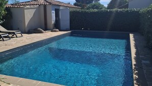 Une piscine rectangulaire bordée de carreaux de pierre, un petit bâtiment avec un toit en tuiles et un arbuste en arrière-plan.