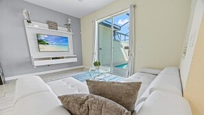 A modern living room with a white sofa, a flat-screen TV mounted on a white wall, a glass table with a small plant, and a sliding glass door leading to a pool area. - Last Minute Disney Getaway! Private Pool & Themed Bedrooms. Book Now! #22516 (Orlando)
