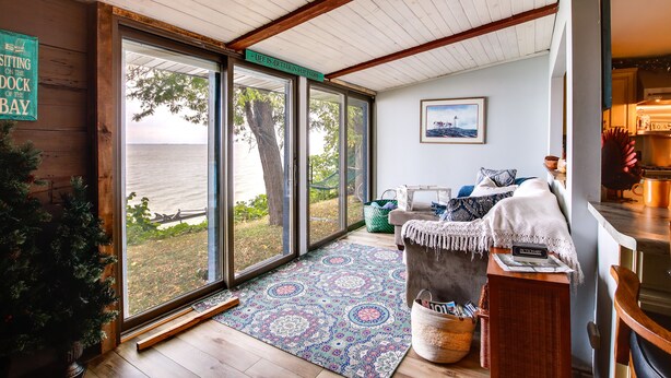 Ein gemütliches Zimmer mit einem großen Schiebeglastür, die zu einem Steg führt, einer Holzbalkendecke, einem gemusterten Teppich, einer Couch mit Kissen und einem durch die Tür sichtbaren Küchenbereich.
