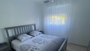 Une chambre à coucher avec un lit, une table de chevet, une fenêtre avec des rideaux et un luminaire suspendu.