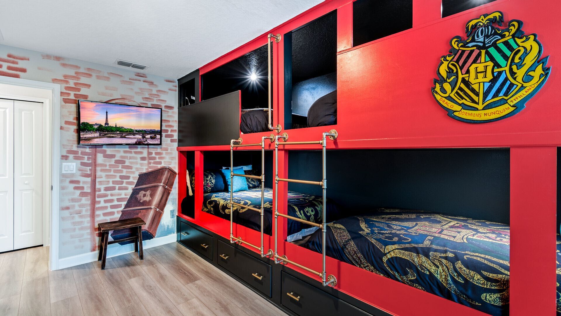 A dorm-style room with a red bunk bed, a flat-screen TV, and a brick-patterned wall.