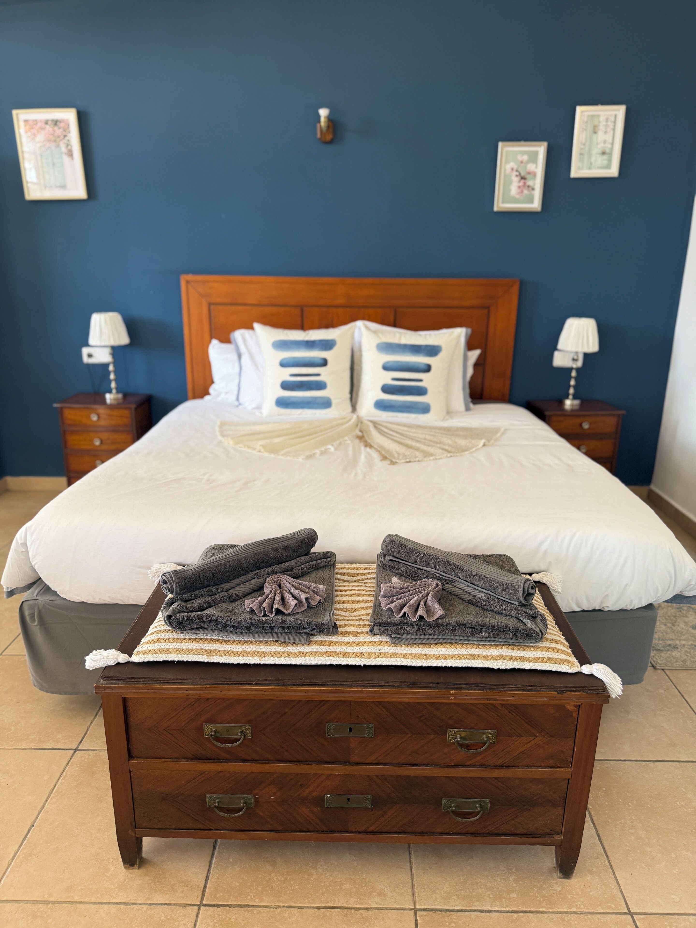 Una cama bien hecha con almohadas blancas y azules, un cabecero de madera y un pequeño aparador de madera al frente.