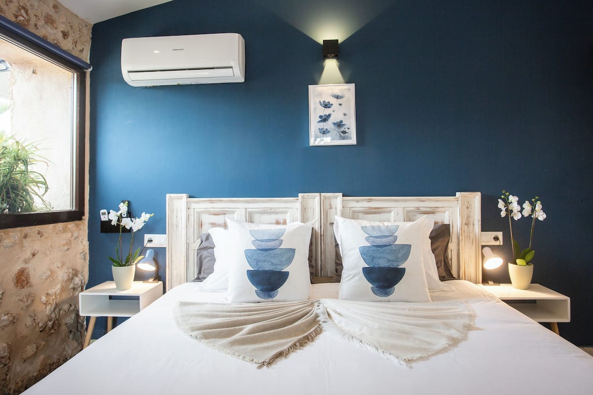 A bedroom with a bed, two nightstands, a wall-mounted air conditioner, a framed picture, and a window with a plant.