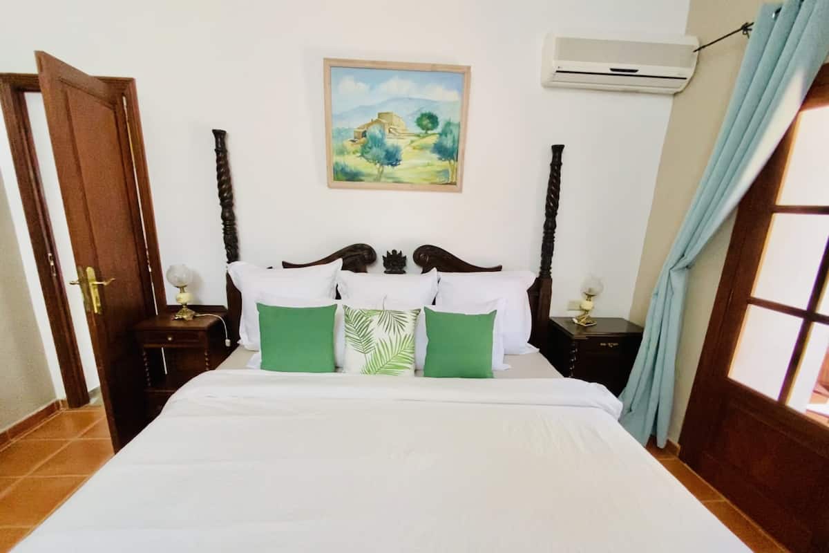 A bedroom with a wooden bed, green and white pillows, a painting on the wall, an air conditioner, and a window with curtains.