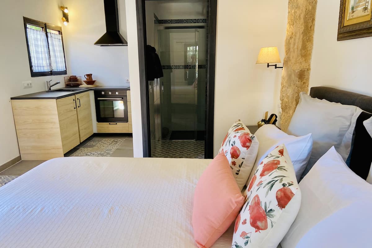 A hotel room with a bed, a nightstand, a lamp, a window with curtains, a kitchen area, and a bathroom.