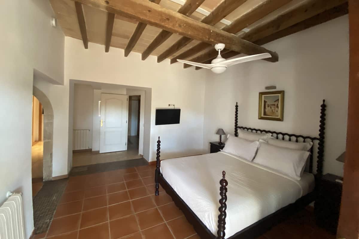 A bedroom with a wooden ceiling, a bed with a dark wooden frame, a white wall, and a tiled floor.