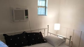 Una cama individual con estructura metálica, una mesita de noche, un aire acondicionado instalado en la pared, una ventana con cortinas y una lámpara de noche pequeña.