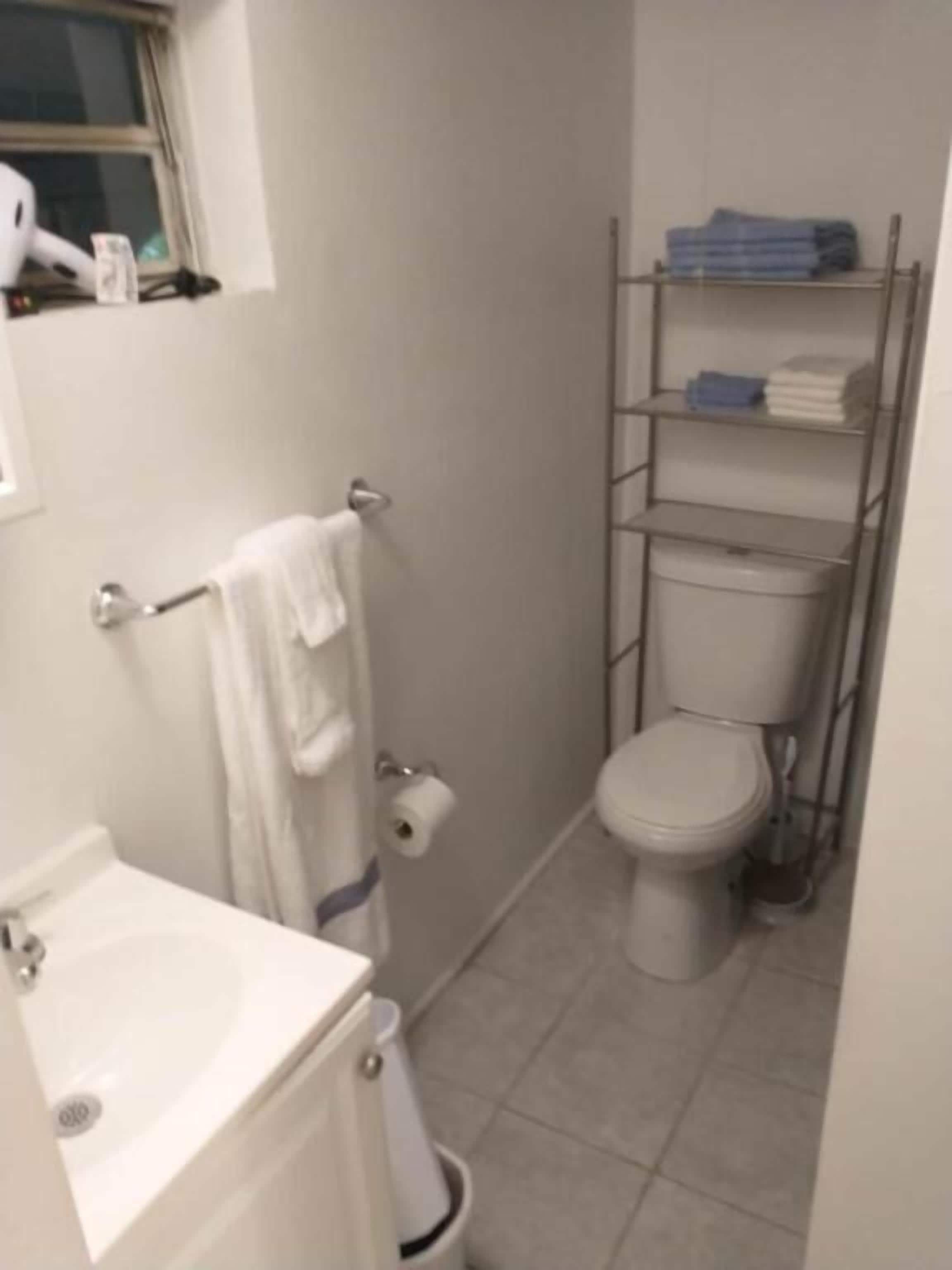 Un baño con inodoro, lavabo, toallero, estante con toallas dobladas y dispensador de papel higiénico.