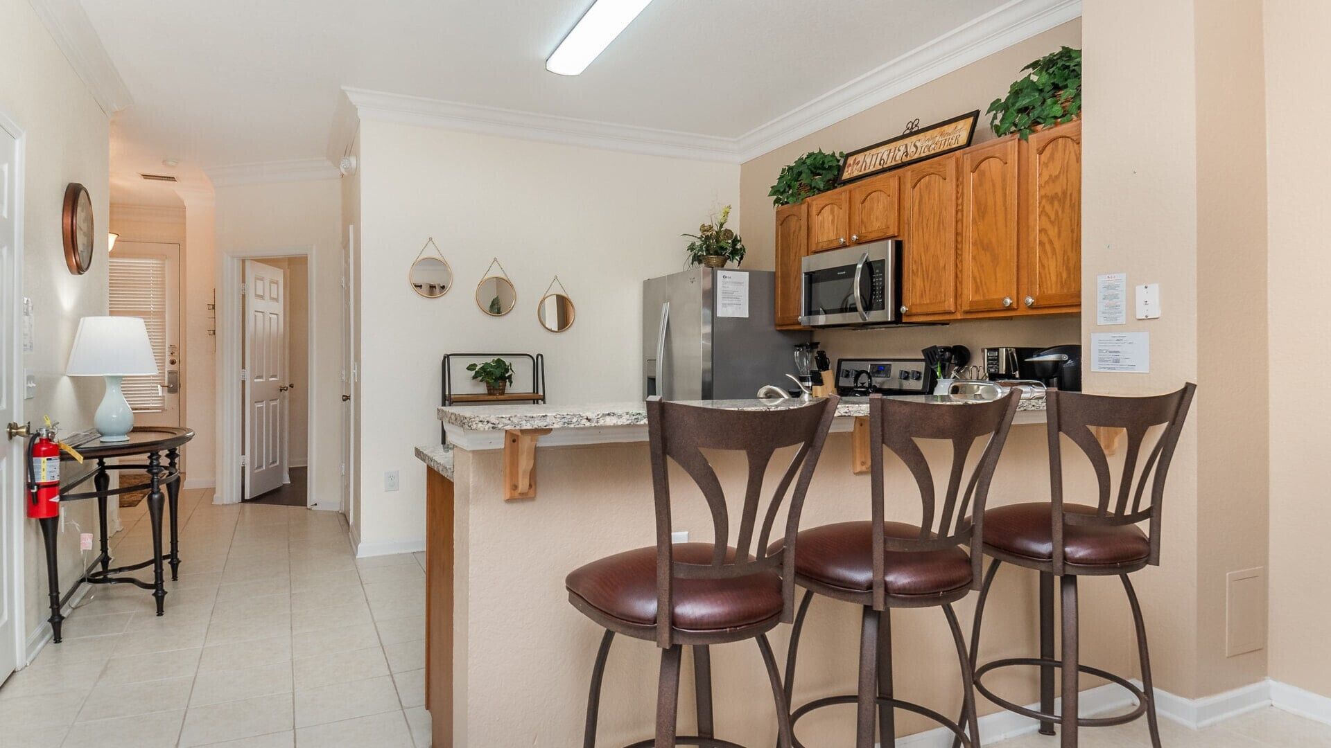 A kitchen with wooden cabinets, a microwave, and a refrigerator. There are three bar stools and a small table with a lamp.