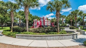 Children can play on the central play structure surrounded by a low wall and palm trees.