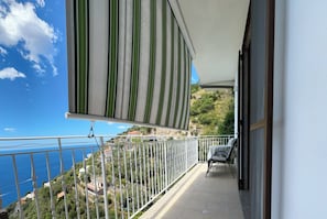 A balcony with a view of the sea and hills, a striped awning, and a table with chairs.