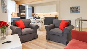 Un salón moderno con dos sillones grises, una mesa blanca y un jarrón con rosas rojas. Un control remoto está sobre la mesa.