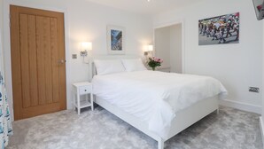 Un dormitorio con una cama grande, una puerta de madera, una mesita de noche, una lámpara de pared, un cuadro enmarcado y un jarrón pequeño con flores.