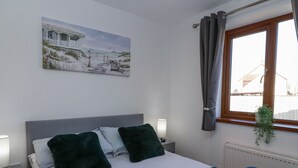 A bedroom with a bed, two pillows, a bedside lamp, a window with curtains, a radiator, and a painting on the wall. - Bluebell Lodge, Meadow View Lodges (Burnham-on-Sea)