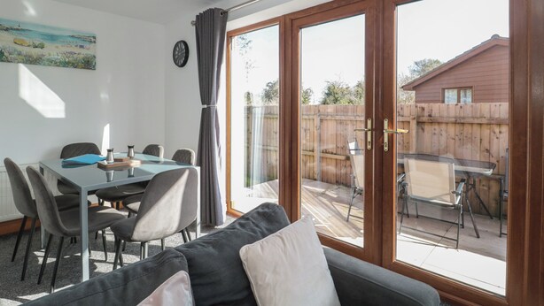 A dining area with a table and chairs, a sofa, and a large sliding glass door leading to an outdoor patio. - Bluebell Lodge, Meadow View Lodges (Burnham-on-Sea)