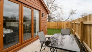 A patio with a glass door, a table, and two chairs. - Bluebell Lodge, Meadow View Lodges (Burnham-on-Sea)