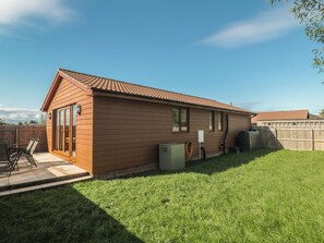 A single-story wooden house with a tiled roof, a fenced backyard, and a patio with outdoor furniture. - Bluebell Lodge, Meadow View Lodges (Burnham-on-Sea)