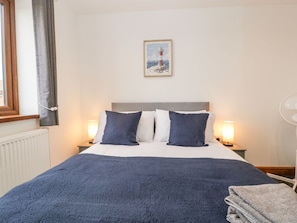 Un lit bien fait, recouvert d’une couverture bleue, avec deux oreillers blancs et deux oreillers bleu foncé. Un tableau encadré représentant un phare est accroché au mur au-dessus du lit.
