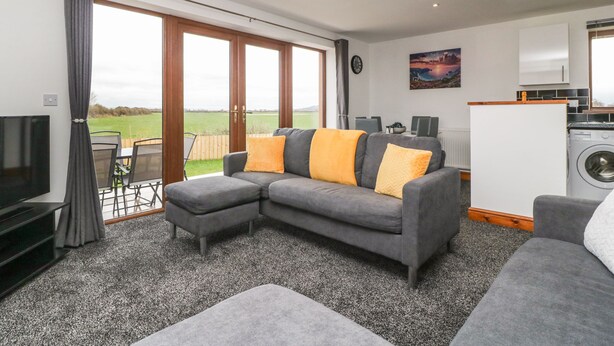 A modern living room with a grey sectional sofa, a flat-screen TV, and a dining area with a table and chairs. - Delphine Lodge, Meadow View Lodges (Burnham-on-Sea)