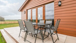 A patio with a glass table and four chairs, a wooden house with large windows, and a view of the countryside. - Delphine Lodge, Meadow View Lodges (Burnham-on-Sea)