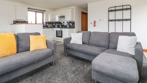 A modern living room with two grey sofas, one with a yellow pillow, and a small white sofa. There is a kitchen in the background with white cabinets and a black countertop. - Delphine Lodge, Meadow View Lodges (Burnham-on-Sea)