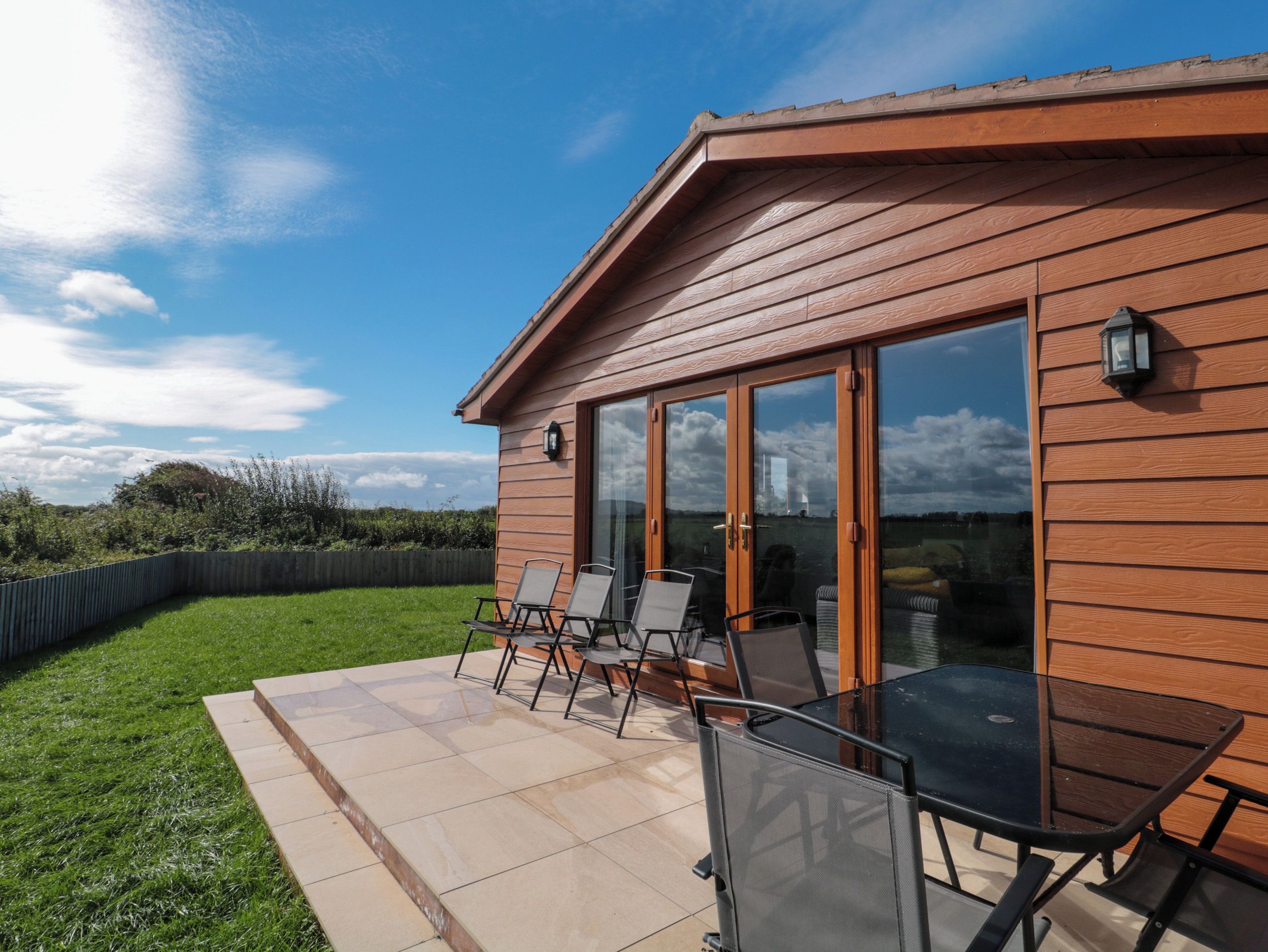A wooden building with a glass door and windows, a patio with chairs and a table, and a grassy area.