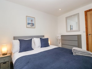 A neatly made bed with a blue blanket and pillows, a bedside lamp, a wall-mounted fan, and a framed picture of a lighthouse.