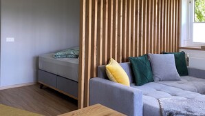 Una sala de estar moderna con un separador de madera, un sofá gris con cojines, una mesa de madera y una cama al fondo.