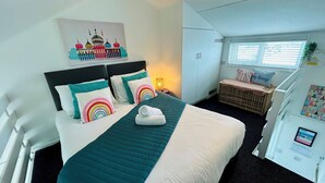 Une chambre d’hôtel comprenant un lit, une table de chevet, une fenêtre avec stores, un climatiseur fixé au mur et un petit coin salon avec un banc et des coussins.