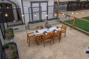 A patio with a wooden table set for four, surrounded by chairs, and a string of lights above. - Private Deck + Firepit + Cornhole | Downtown Paso | Hideaway on Oak (Paso Robles)